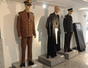 Imagem mostra um dos espaços do Memorial da Justiça Militar de Minas Gerais, com três manequins vestidos com fardamento militar da Polícia Militar de Minas Gerais, do Corpo de Bombeiros de Minas Gerais e uma toga de magistrado civil. As roupas são usadas por magistrados militares e civis durante as audiências na Justiça Militar de Minas Gerais