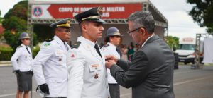 A foto mostra bombeiros com farda branca e o desembargador do TJMMG de terno cinza. Ele coloca uma medalha no aspirante Rodolfo Finamor Costa Gomes