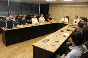 A imagem mostra homens e mulheres sentados em uma mesa de reunião de madeira com detalhes pretos. Eles olham na direção do chefe de gabinete da Presidência, que veste camisa branca e gravata vermelha. Ao fundo há outro grupo de homens e mulheres sentados diante de uma janela com cortinas fechadas