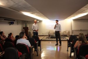A imagem mostra dois homens no auditório do TJMMG diante de uma plateia. À direita está o desembargador Fernando Armando Ribeiro, de camisa clara, calça e sapatos escuros. Ele segura um microfone. À esquerda, o servidor Gustavo Cândido da Silva, de camisa listrada e calça jeans. Ao fundo estão localizadas duas poltronas brancas, uma mesa de vidro, um quadro branco, um púlpito de madeira e três bandeiras