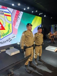 A imagem mostra dois homens lado a lado, em um palco. O homem mais à esquerda da imagem é o coronel Erlando Ferreira da Silva, novo comandante geral da 2ª RPM, e o homem à direita é coronel Flávio Godinho Pereira, que deixou o comando. Eles usam farda da PMMG