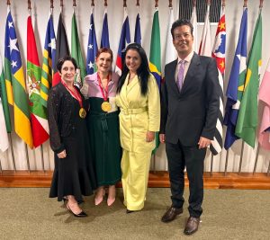 A imagem mostra três mulheres e um homem tendo dispostas as bandeiras de diversos estados brasileiros ao fundo. O homem é o desembargador Fernando José Armando Ribeiro, que veste terno preto, camisa clara e gravata lilás. Ao seu lado está a desembargadora Tânia Regina Reckziegel, presidente do Cojum, que tem cabelos pretos e veste um conjunto de calça e blaser amarelo. No centro das mulheres está a presidente do STM, Maria Elizabeth Guimarães Teixeira Rocha, de vestido verde musgo com casaqueto lilás e sapato rosa. Na outra ponta está a desembargadora Evangelina Castilho Duarte, ouvidora das mulheres do TJMG, de vestido e casaco preto. A ministra e a desembargadora do TJMG exibem uma medalha do Conjum cada, composta por uma moeda dourada presa ao pescoço por uma fita vermelha 