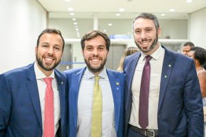A imagem mostra três homens abraçados, que sorriem para a foto. Ao centro está o juiz de Direito da Justiça Militar João Pedro Hoffert Monteiro de Lima, de terno azul, camisa branca e gravata amarela. Do lado esquerdo da imagem está o vice-presidente Jurídico-Legislativo da Adep-MG, Bruno Fiorin Hering, de terno azul, camisa branca e gravata vermelha, e do lado direito está o novo presidente da entidade, Rômulo Luis Veloso de Carvalho, de terno azul, camisa branca e gravata vinho
