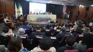 Imagem mostra o auditório do Crea-MG. Em primeiro plano está a plateia, composta por homens e mulheres. No palco há uma mesa com autoridades, bandeiras e um telão ao fundo, músicos no canto esquerdo e um púlpito no lado direito da foto