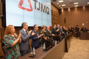 A imagem mostra várias autoridades, entre homens e mulheres, atrás de uma tribuna de madeira. Eles estão de pé aplaudindo. O vice-presidente do TJMMG, desembargador James Ferreira Santos, é o terceiro da esquerda para a direita da imagem. Ele veste terno azul, camisa branca e gravata preta. Na parede, também de madeira, há um telão com a logomarca do TJMG, composta pelo triângulo estilizado de Minas Gerais e as iniciais do nome do órgão em letra maiúscula.