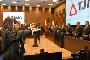 A imagem mostra, no canto esquerdo, os juízes empossados vestindo uma beca preta com a mão direita estendida em posição de juramento. À direita estão oito autoridades que compuseram o dispositivo de honra, sendo seis homens e duas mulheres. As paredes também são de madeira e apresentam um telão com a logomarca do TJMG, composta pelo triângulo estilizado vermelho da bandeira de Minas Gerais e as iniciais do nome do órgão em letra maiúscula.