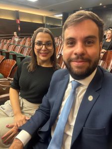 A imagem mostra a juíza usando camisa preta, saia marrom e óculos. Ao seu lado está o juiz vestindo terno azul e gravata azul. Eles estão em um auditório com cadeiras vermelhas e paredes pretas