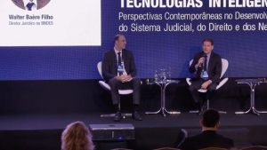 A imagem mostra dois homens sentados diante de um painel. À esquerda da foto está o diretor jurídico  Walter Baère Filho, vestindo terno e gravata preta. À direita está o desembargador Fernando Antônio Nogueira Galvão da Rocha falando ao microfone e vestindo terno e gravata preta. Ao fundo há um painel azul com o nome do palestrante e do congresso 