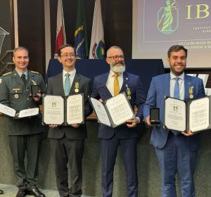 A foto mostra 5 homens em pé, quatro deles seguram o certificado e a medalha de Ordem ao Mérito IBDM. A pessoa mais à direita é o juiz João Pedro Hoffert Monteiro de Lima, vestindo terno e gravata azul. Ao fundo há três bandeiras e um telão com a logo do IBDM