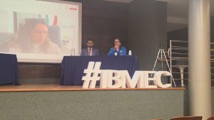 A imagem mostra o palco do Auditório do IBMEC. Há um homem e uma mulher sentados atrás de uma mesa com um pano azul. A frente da mesa há um letreiro com as iniciais do IBMEC. A mulher é a juíza Carolina Aleixo Benetti de Oliveira Rodrigues falando ao microfone, usando camisa azul e óculos. As paredes ao fundo são escuras e há um telão com a transmissão ao vivo da promotora de Justiça Caroline Piloni, vestindo camisa branca e com cabelos abaixo do ombro