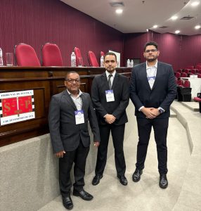 A imagem mostra três homens de pé no auditório do TJSP. À esquerda está o servidor Edivaldo Pereira dos Santos vestindo terno cinza e usando óculos. Ao meio, o juiz George Walter Barreto Paviotti, com as mãos cruzadas à frente do corpo, vestindo terno e gravata pretos. À direita, o servidor Pedro Henrique Capila de Abreu, também com as mãos cruzadas à frente do corpo, usando terno preto e óculos. Eles estão diante de uma tribuna de madeira com a logo do TJSP. Ao fundo há uma parede e cadeiras na cor vinho, enquanto o teto e o chão são brancos