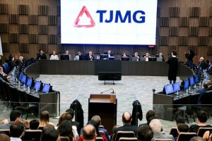 A imagem mostra dez autoridades sentadas em uma tribuna de madeira na sala do Órgão Especial do TJMG, sendo quatro mulheres e seis homens. O primeiro à esquerda da imagem é o ouvidor do TJMMG, desembargador Fernando José Armando Ribeiro. Em primeiro plano estão os convidados da solenidade. Ao fundo há uma parede de madeira e um telão com a logomarca do TJMG, composta pelo triângulo estilizado da bandeira de Minas Gerais e as iniciais em letra maiúscula.  