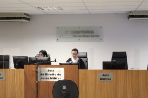 A imagem mostra a juíza Daniela de Freitas Marques vestindo toga e usando óculos. Ela está sentada atrás de uma tribuna de madeira com o letreiro escrito Juiz de Direito do Juízo Militar. Ao seu lado há uma servidora da auditoria ao telefone e acima há o letreiro escrito 3ª Auditoria Justiça Militar de Minas Gerais