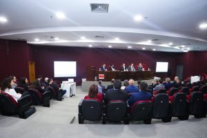 A foto mostra o auditório do TJSP durante a 2ª Reunião Nacional de Gestão de Demandas do Eproc. As paredes ao fundo são vermelhas, o teto e o chão são brancos. Os participantes estão sentados em cadeiras na cor vinho e há cinco homens em posição de destaque sentados na tribuna de madeira