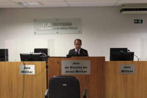 A foto mostra o juiz de Direito Marcelo Adriano Menacho dos Anjos usando terno preto, gravata vermelha e óculos. Ele está atrás de uma bancada de madeira com o letreiro de Juiz de Direito do Juízo Militar. A parede ao fundo é branca e apresenta uma placa onde está escrito 1ª Auditoria Justiça Militar de Minas Gerais