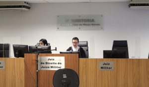 A imagem mostra a juíza Daniela de Freitas Marques vestindo toga e usando óculos. Ela está sentada atrás de uma tribuna de madeira com o letreiro escrito Juiz de Direito do Juízo Militar. Ao seu lado há uma servidora da auditoria ao telefone e acima há o letreiro escrito 3ª Auditoria Justiça Militar de Minas Gerais