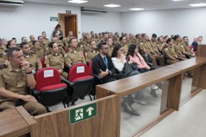 Na imagem estão alunos do Curso de Habilitação de Oficiais da PMMG, fardados, e alguns civis dispostos na sala de audiência do Tribunal Pleno do TJMMG, com cadeiras vermelhas e paredes brancas.