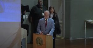 A imagem mostra o desembargador Rúbio Paulino Coelho falando em uma tribuna de madeira, ao microfone. Ele veste terno cinza e gravata vermelha