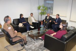 A foto mostra quatro representantes do TJMMG em encontro com desembargador Antônio Gomes de Vasconcelos do TRT-MG. Todos estão sentados em poltronas pretas em volta de uma mesa de vidro. À esquerda está o desembargador James Ferreira Santos usando farda oficial da PMMG. Ao fundo está a chefe do Gabinete do NCJ, usando um conjunto de blusa e calça azul marinho, acompanhado por botas brancas. Ao seu lado está o desembargador do TRT- MG usando calças pretas e blazer marrom. À direita está o desembargador Jadir Silva vestindo terno preto e gravata amarela, e o desembargador Fernando Antônio Nogueira Galvão da Rocha vestindo terno e gravata azul. Em primeiro plano está juíza Carolina Aleixo Benetti usando um conjunto de calça e blusa vermelha. As paredes da sala são brancas e no chão há um tapete de tons terrosos