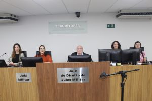 A imagem mostra 5 pessoas atrás de uma mesa de madeira com computadores. Ao centro está o juiz João Libério da Cunha usando terno preto e óculos. À esquerda está a juíza Carolina Aleixo Benetti de Oliveira Rodrigues usando blusa vermelha e óculos. À direita está a servidora Bárbara Marques sorrindo. Na extremidade esquerda está a gerente da 2ª AJME Nádia Prata Neves vestindo camisa preta e casaco listrado e na extremidade direita está a servidora Vanessa Graziela usando blusa rosa
