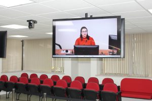 Na imagem mostra uma sala de paredes brancas, com cortinas claras, bancos vermelhos e pretos e com duas televisões em posições inversas. A que está de frente para a imagem mostra a Juíza de Direito Substituta do Juízo Militar Carolina Aleixo Benetti de Oliveira Rodrigues, que está usando uma blusa vermelha de gola alta, óculos pretos e tem cabelos pretos na altura dos ombros