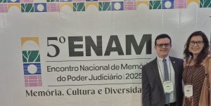A imagem mostra um homem e uma mulher posando para foto em frente a um painel escrito 5º Enam. O homem é o juiz Bruno Cortez Torres Castelo Branco usando terno cinza, gravata azul e óculos. Ao seu lado está a servidora Maria Luiza Silveira Faria usando um vestido marrom com listras brancas, uma bolsa marrom e óculos