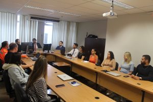 A imagem mostra treze pessoas entre homens e mulheres no plenário do TJMMG sentados atrás de uma mesa de madeira comprida em formato oval. Na extremidade da esquerda está a secretária da Corregedoria, Gislene Amarantes Cunha, usando camisa com estampa zebrada. Mais ao fundo está o coronel João Guilherme Brito Vieira, corregedor do CBMMG, usando farda oficial dos bombeiros. Na cabeceira da mesa está o juiz Marcos Luiz Nery Filho vestindo terno preto e gravata vermelha, olhando para um computador. A esquerda está o juiz George Walter Barreto Paviotti vestindo terno e gravata azuis