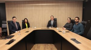 A imagem mostra três homens e duas mulheres em uma mesa de reunião na Sala Diamante do TJMMG. A mesa, assim como o painel por trás deles, é em tons de madeira e preto. O homem mais à esquerda é o juiz auxiliar da Corregedoria do TJMG, de terno preto, camisa branca e gravata vinho com estampa. Ao seu lado está a desembargadora do TJMG, de blaser preto e blusa amarela. Ao centro está o vice-presidente do TJMMG, de terno preto, camisa branca e gravata azul. Ao seu lado está a juíza de Direito do Juízo Militar, de blusa preta, e o assessor de juiz, de camisa azul