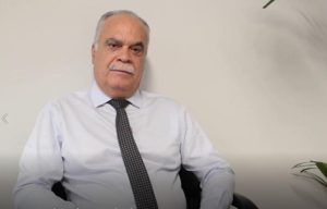 A imagem mostra o desembargador Rúbio Paulino Coelho sentado, de camisa branca e gravata preta com detalhes brancos. Ele tem bigode branco e semblante sério. Ao fundo há uma parede branca com algumas plantas