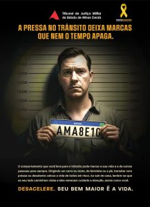 A imagem reproduz o cartaz oficial da campanha Maio Amarelo, com o texto “A pressa no trânsito deixa marcas que nem o tempo apaga. O comportamento que você leva para o trânsito pode marcar a sua vida e a de outras pessoas para sempre. Dirigindo um carro ou moto, de bicicleta ou a pé, transitar com pressa ou desatento coloca a vida de todos em risco. Ao sair de casa, lembre-se que ao seu lado caminham vidas e elas merecem cuidado e atenção, assim como você”. Em destaque na última linha, há o slogan “Desacelere. Seu bem maior é a vida”. Na parte superior do cartaz está a logomarca do TJMMG, composta pelo triângulo vermelho estilizado da bandeira de Minas Gerais com o nome do órgão por extenso. Ao lado há a logomarca da campanha, que inclui uma fita amarela e o nome Maio Amarelo. Ao centro há a imagem de um homem branco posando para foto em frente a um painel de delegacia, antes de ir preso. Ele apresenta feições de arrependimento, veste camisa marrom e segura uma placa de carro com as letras em alusão à palavra “amarelo”, a cor da campanha