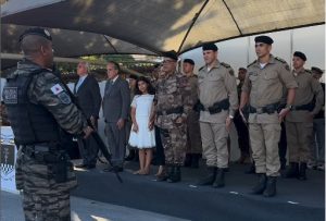 A imagem mostra vários integrantes da PMMG vestindo farda oficial, dentre outros convidados do evento, todos em posição de respeito. Na primeira fileira há seis pessoas, sendo cinco homens e uma garota usando um vestido branco. O segundo da esquerda para a direita é o vice-presidente do TJMMG, desembargador coronel James Ferreira Santos, usando terno e gravata cinzas. No lado direito da imagem está o comandante-geral da PMMG, coronel Frederico Otoni Garcia. Eles estão em cima de um tablado coberto olhando para um integrante da PMMG que segura um armamento