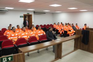 A imagem mostra uma turma de bombeiros militares vestindo farda laranja da corporação. Eles estão sentados na plateia da sala de audiências do Tribunal Pleno do TJMMG, composta por cadeiras vermelhas. Na primeira fileira está o uiz de Direito do Juízo Militar Marcos Luiz Nery Filho, de terno escuro