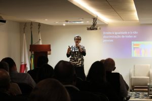 A foto mostra a professora Andréa de Campos Vasconcellos, ao centro da imagem, com uma blusa branca com listras pretas e cabelo curto grisalho. Ela está diante de um slide onde está escrito “Direito à igualdade e à não discriminação de todas as pessoas”. A palestrante está no auditório do TJMMG, em frente à plateia