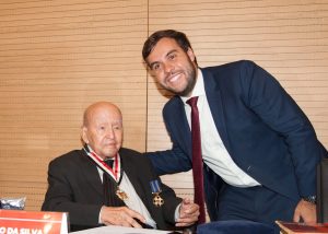 A imagem mostra o momento em que o juiz João Pedro Hoffert Monteiro de Lima entrega a Medalha do Mérito Judiciário Militar ao professor José Afonso da Silva. O homenageado está sentado, veste terno e gravata pretos. O juiz veste terno azul escuro com gravata vermelha. Eles estão atrás de uma mesa preta com uma placa com o nome do professor. A parede ao fundo é de madeira