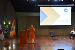 A imagem mostra a juíza Carolina Aleixo Benetti de Oliveira Rodrigues durante palestra no CBMMG. Ela está apoiada ao púlpito de madeira falando ao microfone, tem cabelos pretos na altura dos ombros, e usa um vestido escuro e sapatos pretos. O chão é de madeira e a parede é cinza. Ao fundo há um telão exibindo o brasão da CBMMG e informações sobre o Seminário. Ao lado da magistrada estão três bandeiras: de Minas Gerais, do Brasil e do CBMMG 