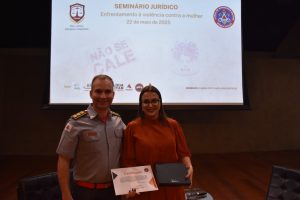 Na imagem está a juíza Carolina Aleixo Benetti de Oliveira Rodrigues e o Corregedor do CBMMG, coronel João Guilherme Brito Vieira. A juíza segura uma caixa preta na mão direita e na esquerda um certificado do Seminário Jurídico. Ela usa roupas escuras, óculos pretos e sorri para a foto, já o coronel está de farda do Corpo de Bombeiros. Ao fundo há um telão exibindo o brasão da CBMMG e informações sobre o Seminário