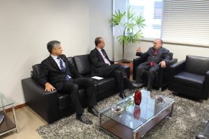 A imagem mostra três homens sentados no gabinete da Presidência do TJMMG, em um sofá preto. A sala possui paredes brancas, piso claro, um tapete cinza e uma mesa de centro, com um vaso de plantas próximo à janela. O primeiro à esquerda da foto é o vice-presidente eleito do TRE-MG, desembargador Carlos Henrique Perpétuo Braga, vestindo terno preto e gravata azul. A sua esquerda está o presidente eleito do TRE-MG, desembargador Júlio César Lorens, com terno preto e gravata preta. O terceiro é o presidente do TJMMG, desembargador Jadir Silva, que veste terno preto e gravata vermelha.