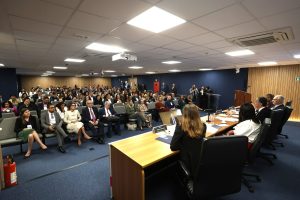 A imagem mostra o auditório do Conselho Nacional de Justiça (CNJ), que possui carpete azul-escuro no chão e paredes com partes em madeira ripada clara e partes em carpete azul-escuro. Em primeiro plano, há uma mesa comprida de madeira clara com cinco pessoas sentadas em cadeiras pretas, sendo elas os palestrantes, todos vestidos com roupas sociais. Em segundo plano, há várias pessoas sentadas em cadeiras cinzas e pretas, também usando roupas sociais.