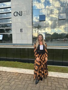 Na imagem mostra a Gerente da Assessoria de Comunicação Institucional do TJMMG Tatiana Reis, usando um vestido longo de manga comprida marrom, com círculos preto e laranja, um colete preto e cinto preto, em frente ao edifício-sede do CNJ, em Brasília, um edifício com fachada de vidro e concreto cinza.