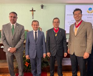 Na imagem estão quatro homens posando para a foto em uma sala que, na parede ao fundo, há uma cruz e uma tela com informações do evento. Dois homens usam medalhas penduradas no pescoço, são eles, o desembargador Fernando José Armando Ribeiro, que usa um blazer bege e calças escuras; e o desembargador José Marcos Vieira do TJMG que usa um terno preto. Os homens que estão sem medalhas são o desembargador Estevão Lucchesi, que usa terno cinza com gravata verde; e o presidente do TJMG Luiz Carlos Corrêa Junior, que usa terno azul marinho e blusa branca e o presidente do TJMG, desembargador Luiz Carlos Corrêa Junior, que usa terno azul marinho e blusa branca