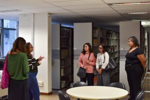 A imagem mostra a Biblioteca do TJMMG durante visita guiada de estudantes de Direito da Universidade Fumec. Há cinco mulheres, sendo que à esquerda estão duas servidoras do Tribunal orientando as três estudantes que estão à esquerda. A primeira servidora segura uma bolsa rosa e veste blusa verde e saia preta. A segunda usa blusa listrada e calça jeans. A primeira estudante está segurando uma bolsa preta e usa blusa rosa com calça preta. A segunda usa blusa com casaco brancos e calça preta, ela também segura uma blusa preta. A terceira está usando camisa e calça pretas. Ao fundo há uma prateleira com vários livros e em primeiro plano há uma mesa redonda com quatro cadeiras pretas ao redor