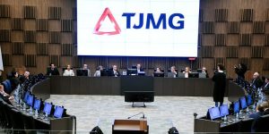 A imagem mostra dez autoridades sentadas em uma tribuna de madeira na sala do Órgão Especial do TJMG, sendo quatro mulheres e seis homens. O primeiro à esquerda da imagem é o ouvidor do TJMMG, desembargador Fernando José Armando Ribeiro. Em primeiro plano estão os convidados da solenidade. Ao fundo há uma parede de madeira e um telão com a logomarca do TJMG, composta pelo triângulo estilizado da bandeira de Minas Gerais e as iniciais em letra maiuscula
