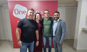 A imagem mostra um grupo de três homens e uma mulher. Eles estão de pé diante de um banner vermelho. O homem mais à direita da imagem é o professor Diocesio Sant'anna da Silva, de terno cinza