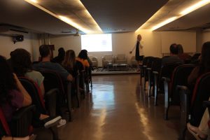 A imagem mostra o Auditório do TJMMG com diversas pessoas sentadas assistindo a palestra da psicóloga Júlia Ramalho, ela está em pé, falando ao microfone, vestindo blazer claro e vestido escuro