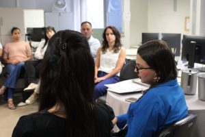 A imagem mostra um grupo de pessoas reunido em uma sala de escritório, sentadas em círculo, participando da roda de conversa sobre o autismo. Em primeiro plano está a juíza Carolina Aleixo Benetti de Oliveira Rodrigues usando blusa azul e óculos. Ao fundo, há computadores, armários e ventiladores