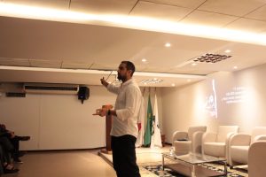 A imagem mostra o nutricionista Pedro Barros realizando uma apresentação no Auditório do TJMMG. Ele veste camisa clara e calça preta. Ao fundo há três cadeiras brancas vazias atrás de uma mesa de centro de vidro em cima de um tapete. As paredes são brancas e há três bandeiras no canto da sala