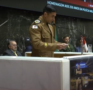 A imagem mostra o comandante-geral da PMMG, coronel Carlos Frederico Otoni Garcia, durante seu discurso. Ele está em pé atrás de um púlpito, falando ao microfone e segurando um papel. Ele usa farda oficial da PMMG. Atrás dele, está o desembargador Jadir Silva usando terno preto, gravata amarela e óculos. A parede é de mármore preto e contém um painel eletrônico exibindo a transmissão ao vivo da solenidade