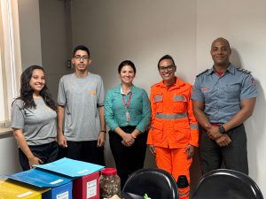 A imagem mostra 5 pessoas, sendo três mulheres e dois homens. Os primeiros três são membros da equipe da Gestão Documental, sendo que dois vestem camisa cinza e calça preta como uniforme e uma veste camisa verde e calça preta. A quarta veste farda laranja oficial da CBMMG e o quinto veste roupa azul oficial da CBMMG. Em primeiro plano há uma mesa com caixas coloridas e atrás há cadeiras pretas. A parede ao fundo é branca