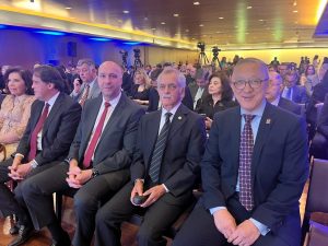 A imagem mostra três desembargadores representando diferentes Tribunais de Justiça Militar, sentados em um auditório. O primeiro é Rodrigo Mohr, do Rio Grande do Sul, vestindo terno preto e gravata vermelha. O segundo é Sócrates Edgard dos Anjos, de Minas Gerais, usando terno preto e gravata listrada. O terceiro, de São Paulo, veste terno azul com gravata e óculos. Ao fundo, vê-se a plateia do evento e algumas câmeras.