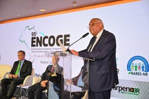 A imagem mostra o presidente do Colégio de Corregedoras e Corregedores-Gerais de Justiça, desembargador Gilberto Barbosa, falando ao microfone em um púlpito de vidro durante o discurso de abertura. Ele segura um papel, usa terno azul com gravata da mesma cor e óculos. Ao fundo, está o painel do 95º Encoge e dois convidados no dispositivo de honra.
